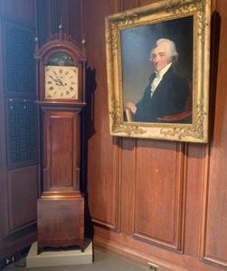 color photograph showing a tall case clock in the corner and a framed portrait of a man on the wall to the left.